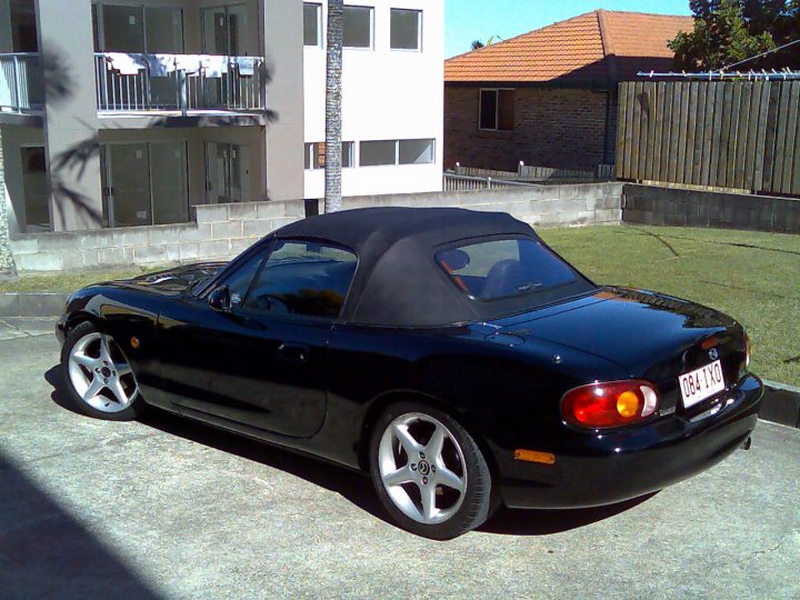 Pistonheads - The image shows a black electric sports car parked on a concrete surface. The vehicle features a convertible top and is equipped with a distinctive rear spoiler and characteristic circular taillights. It is positioned next to a modern apartment building under a clear blue sky, with a neighboring house and wooden fence in the background. The vehicle's bodywork is sleek and aerodynamic, indicative of its sporty design, and the overall setting suggests a suburban residential area.