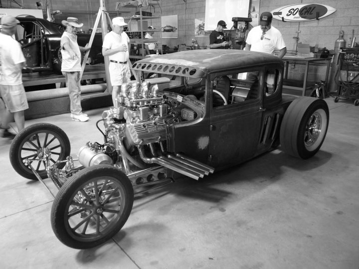 Pistonheads Hot Liking Moment Rodsi - The image appears to be a black and white photograph showing an antique race car, likely from the early to mid-20th century, displayed inside a garage or workshop. The car is predominantly black with chrome accents, featuring an open cockpit and a long wheelbase. Around the vehicle are several individuals, dressed in casual attire, observing the car with interest. The setting suggests a display or exhibition, with the car possibly being the main attraction, and the people might be interested spectators or mechanics.