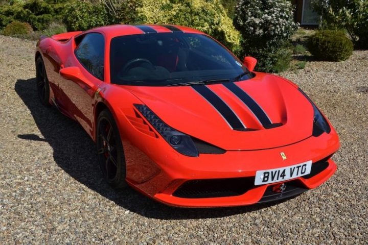 458 Speciale - Page 1 - East Anglia - PistonHeads - The image shows a sleek, red two-door coupe parked on a gravel surface. The car is a sports car, identifiable by its aggressive front spoiler and low, wide stance. It features a double stripe across the hood and a similar stripe pattern repeated on the sides. The headlights are pointed and give the vehicle a powerful look. The vehicle is positioned at an angle to the camera, showing the side profile and part of its front fender. In the background, there is a hint of greenery.