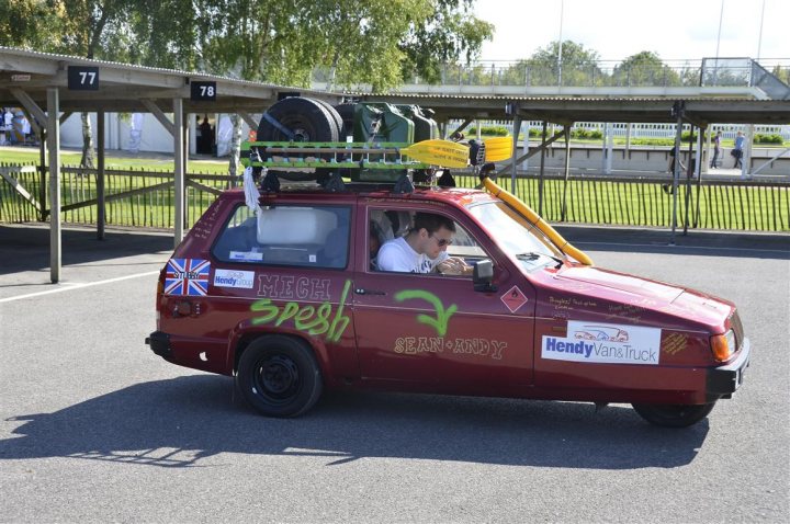 Mongol Rally 2011 - Page 1 - Goodwood Events - PistonHeads - The image depicts a vibrant scene featuring a red SUV adorned with several stickers and symbols, suggesting it may be a promotional vehicle for a business or event. There is plaid fabric, possibly a canopy or protective material, securly strapped to the roof of the vehicle. The vehicle appears to be in motion, as indicated by the dynamic angle and the motion blur of the wheels. A person is seen hanging out of the backseat window, adding to the lively and spontaneous feel of the scene. There are also people visible in the background, possibly indicating that the vehicle is part of a larger gathering or event.