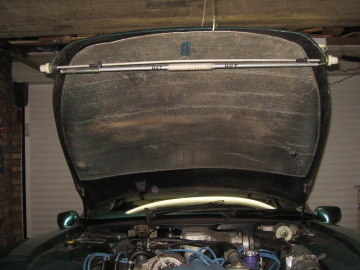 Sneek peak of the new Leven Bonnet stay - Page 1 - Griffith - PistonHeads - The image shows the open trunk of a vehicle. The trunk is propped open by a long metal rod. The interior of the trunk appears dusty and worn. The vehicle, which appears to be an older model due to its undercarriage visible in the photo, has a visible blue tube beneath the engine. The backdrop of the photo is an indoor area, possibly a garage.