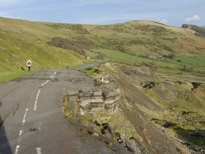 Pistonheads Roads Disusedabandoned - The image showcases a rural landscape where a winding road cuts through the countryside. An individual is captured walking along the side of the road, lending a sense of scale and activity to the scene. The road leads towards a distant valley bordered by rolling hills, which are lush with green grass. Further into the distance, a river or stream meanders through the landscape. The sky above is a clear, blue canvas, providing a serene backdrop to the scene.