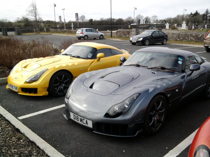 Pistonheads - The image showcases a row of three sport cars parked in a gravel lot. The first two cars are yellow and silver, and the third one is a sleek black coupe. They are all facing the same direction and align with the parking lines. The cars appear to be in good condition and are likely to be of the sports or luxury variety. The lot itself is a gravel surface with patches of grass, and the sky above is filled with clouds.