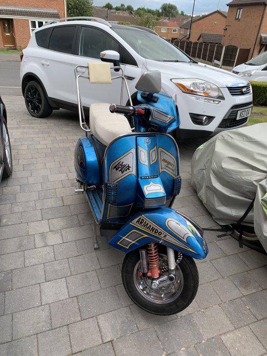 Show us your scooter thread - Page 5 - Biker Banter - PistonHeads UK - The image shows a blue and black moped parked in front of a brick paved driveway. A car is visible behind the moped, partially obscured by its presence. There is a white vehicle to the right of the frame, and a sidewalk can be seen on the left side. In the background, there's a glimpse of a residential area with houses and trees. The setting suggests a quiet suburban neighborhood.