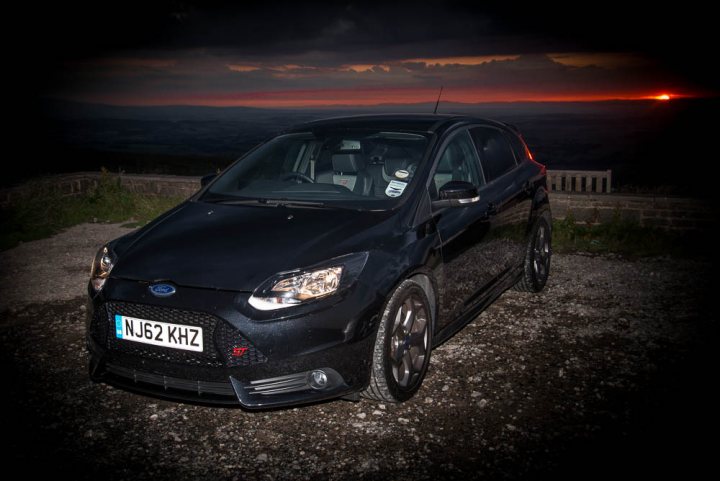 A car parked on the side of a road next to a fire hydrant - Pistonheads - The image captures a black Ford Focus car, parked during twilight. The car is facing the camera, parked on a gravel surface with a hint of a rock garden in the background. The rear of the car is illuminated by a nearby light source, casting a soft glow on the vehicle. In the distance, a dramatic contrast is seen with an orange sun setting behind a mountain, offering a picturesque backdrop to the parked car.