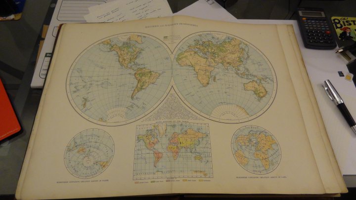 Something different...1895 Times Atlas of the World - Page 1 - Books and Literature - PistonHeads - In the image, an open book is prominently displayed on a desk, showcasing two large world maps. The primary map occupies most of the book, with a smaller inset map situated beneath it. The maps illustrate various aspects of the world, including countries and their locations. In the background, you can see a few personal items like a pocket calendar, a smartphone, and a calculator, indicating the reader's workspace. The desk is cluttered, suggesting an active and perhaps busy environment.