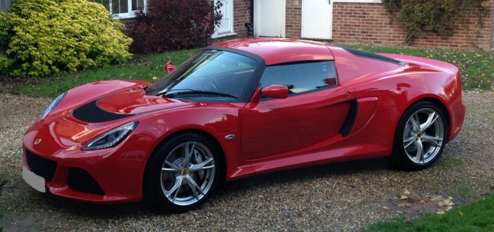 Exige S - Should I? - Page 1 - Elise/Exige/Europa/340R - PistonHeads - The image depicts a striking, sleek red sports car parked on a driveway in front of what appears to be a house or apartment building. The vehicle is impeccably clean and well-kept, with a low profile and vibrant, glossy paintwork. The car suggests high performance and state-of-the-art engineering, with aesthetic features that add to its aerodynamic design. The car's contrasting gray roof, visible through the windshield, provides a nice balance to its bold red body. The setting appears to be a residential area, and there is a sense of tranquility to the scene.