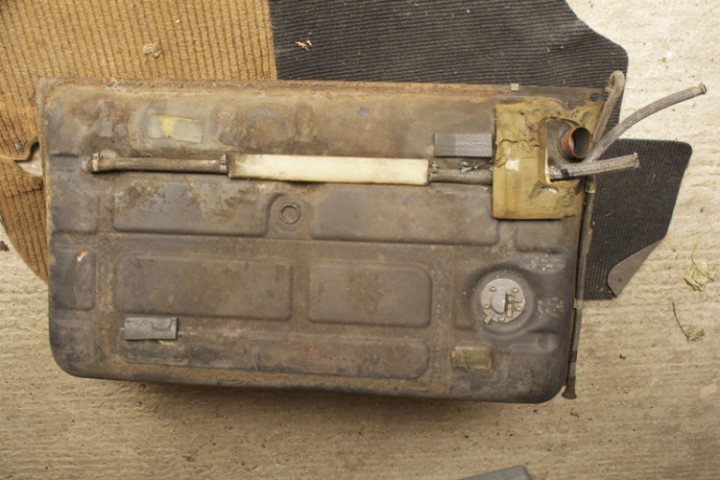 Fitting a 924 Turbo tank to a 944 S1? - Page 1 - Front Engined Porsches - PistonHeads - The image shows an old, heavily worn and dirty car battery series. The battery has a visible label with the numbers "045". Various labels and symbols such as logos, charts, and written texts are partially visible and appear to be faded. The battery is slightly upside down, revealing a thick white strap, and a single metal bolt into which a wooden dowel has been inserted. The battery is placed on a carpeted floor which appears to be an upholstery service setting.