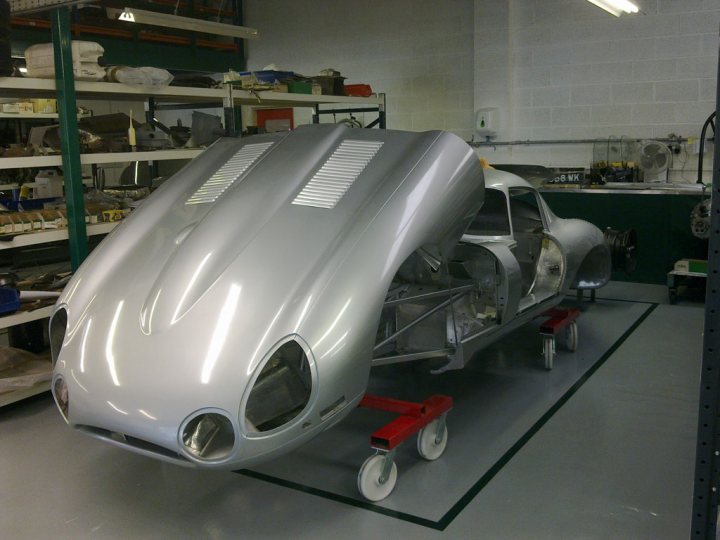 Pistonheads - The image depicts a classic silver racing car resting on a pair of red and black dolly wheels, suggesting it's in motion or being maneuvered in a workshop or garage. This setting indicates that the car may be undergoing restoration or maintenance. The interior of the car is visible, revealing a complex mechanical design. The environment appears industrial, with shelves containing various parts and tools behind the car, and green walls and a metal frame forming part of the car's display platform.