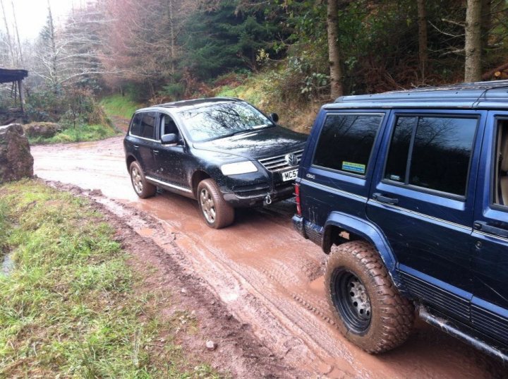 cleaning the off roader - Page 1 - Off Road - PistonHeads - The image shows an off-road situation where a dark-colored SUV is stuck in mud on a trail. A lighter-colored SUV is seen nearby, possibly attempting to assist. The second vehicle's doors are open, suggesting that the driver or passenger might be trying to help. The scene is outdoors, with trees in the background, casting shadows on the vehicles. The ground appears wet and muddy, implying that recent rain or water activity has led to this situation.