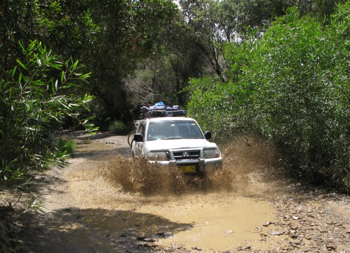 Sydney January Run - Page 1 - Australia - PistonHeads - In the image, a white SUV is driving down a muddy road with trees and lush vegetation on either side. The car appears to be navigating through a splash of brown water. The terrain is slippery and challenging, suggesting that it might have rained recently. The dense foliage indicates a tropical or subtropical environment. The car's movement is kicking up mud from the road.