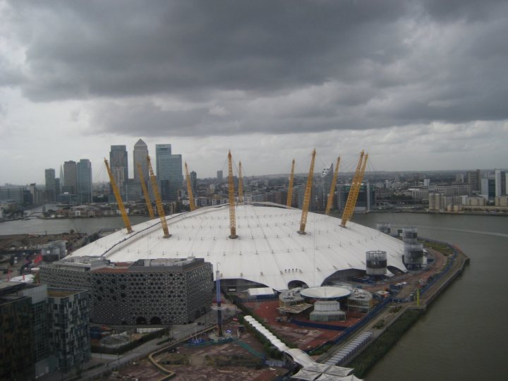 Cable car across London - Page 1 - Boats, Planes & Trains - PistonHeads - The image showcases the distinctive, large dome of the O2 Arena, often referred to as the Millennium Dome. The arena is surrounded by a wetland area where water appears to be separating the main land from a crane-filled construction or industrial site. In the background, the city shows an array of skyscrapers and buildings, hinting at a densely populated urban area. A bridge is partially visible over the wetland. The sky is overcast and gray, suggesting cloudy weather.