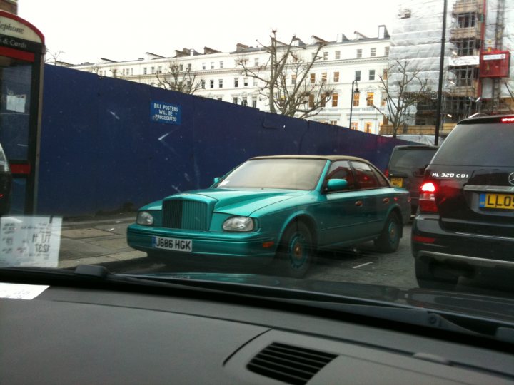 Identify Pistonheads - The image captures a scene on a busy street. A bright blue Bentley car is the prominent vehicle, driving off underneath a clear blue sky. The Bentley's unique design sets it apart from the other vehicles on the road, which include several cars of various colors and models. The street is lined with a mix of buildings and what appears to be a large blue structure. The perspective of the image suggests it might be taken from inside a car on the street, adding a sense of spontaneity and urban life.