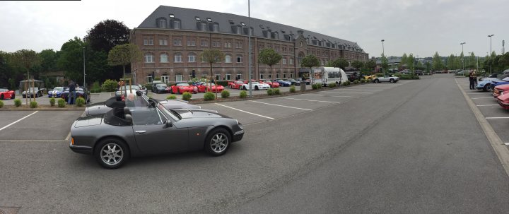 A car driving down a street next to tall buildings - Pistonheads - The image depicts a vast parking lot, primarily occupied by numerous red sports cars. These cars are scattered around the lot, some parked closer to the camera, while others are further away. The lot is a broad, open space, with a road running through the middle, meandering from the foreground to the distance.

Dominating the background is a large, red-bricked building, characterized by its multiple floors and sloped roof. The building is flanked by a mix of trees and bushes, adding a touch of greenery to the otherwise urban setting. On the right side of the image, a few additional vehicles can be seen. These include a bus and a truck, adding variety to the parking lot's composition. The overall scene suggests a vibrant atmosphere, possibly in a bustling city or town where sports cars are a common sight.