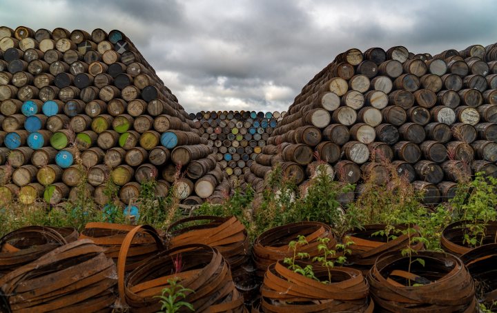 Show us your whisky! Vol 2 - Page 260 - Food, Drink & Restaurants - PistonHeads UK - The image presents a scene of a large stack of wooden barrels in an outdoor setting. These barrels, varying in size and color, are piled up haphazardly, with some stacked atop each other. In the background, there's a field with a few scattered items, including what appears to be a small metal structure. The sky is overcast, adding a somewhat moody atmosphere to the scene.