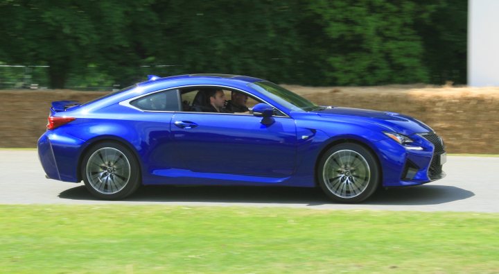 Goodwood moving motor show photos - Page 1 - Goodwood Events - PistonHeads - The image captures a vibrant blue Lexus coupe in motion on a road. The car is well-lit, suggesting it's daytime. Inside the car, you can see three individuals enjoying the ride. The surroundings include green grass and a dirt partition on one side, indicating a rural or semi-rural area. The overall atmosphere of the image is lively and dynamic, reflecting the energy of the moving car and its occupants.