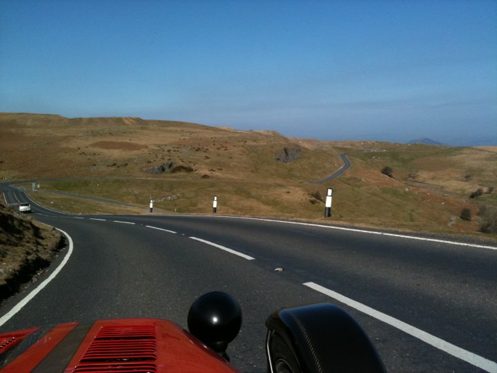 Pistonheads Wales Blat - The image presents a picturesque scene from inside a car. The perspective is one of a passenger seated in the backseat, with the car winding its way along a curvy road situated in a landscape of rolling hills and a distant mountain. The road is bordered by lush greenery on the left, and on the right, there are guardrails and occasional directional signs for drivers. The sky above is a clear blue, suggesting it is a bright day.