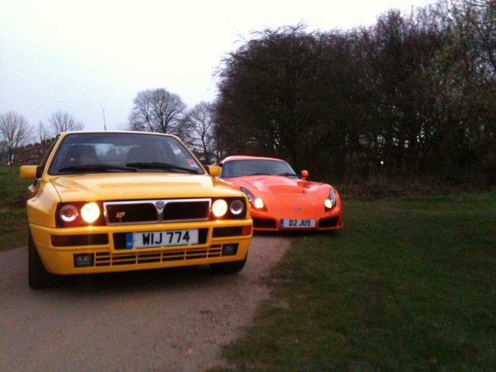 Pistonheads Seconds Endulge - The image shows two sports cars parked on a gravel pathway. One car is bright yellow with white headlights and a license plate that reads "WIJ 774." The second car is vibrant orange and is adorned with a convertible top. The vehicles are set against a backdrop of trees and grass, illustrating a serene outdoor setting, possibly during dawn or dusk given the soft, diffused lighting in the sky. The orange sports car stands out against the gravel road, creating a striking contrast.