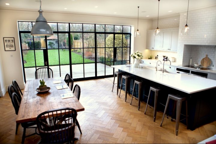 Edwardian semi - New extension and refurb - Page 1 - Homes, Gardens and DIY - PistonHeads - The image portrays a modern open-concept kitchen and dining area within a residential setting. A large wooden dining table is situated in the center with black chairs, placed at each spot. Off to the side, a central island countertop with stools for seating is adorned with a contrast of black and white colors. The kitchen boasts white cabinetry, sleek stainless steel appliances, and an abundant display of white subway tiles. Large windows and sliding glass doors create a seamless transition to the thriving greenery of a garden beyond, which adds a touch of nature to this contemporary interior.