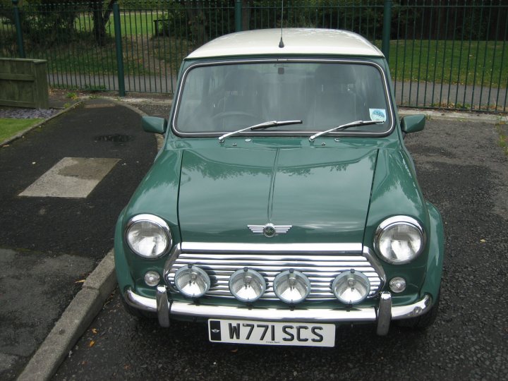 green inspriation - Page 1 - Classic Minis - PistonHeads - The image displays a classic green Mini Cooper model car parked on a street with a blacktop. The car has a vintage design with two round headlights and two round auxiliary lights mounted on the front. On the rear of the vehicle, there is a license plate. The vehicle appears to be well-maintained and is facing towards the right side of the image.