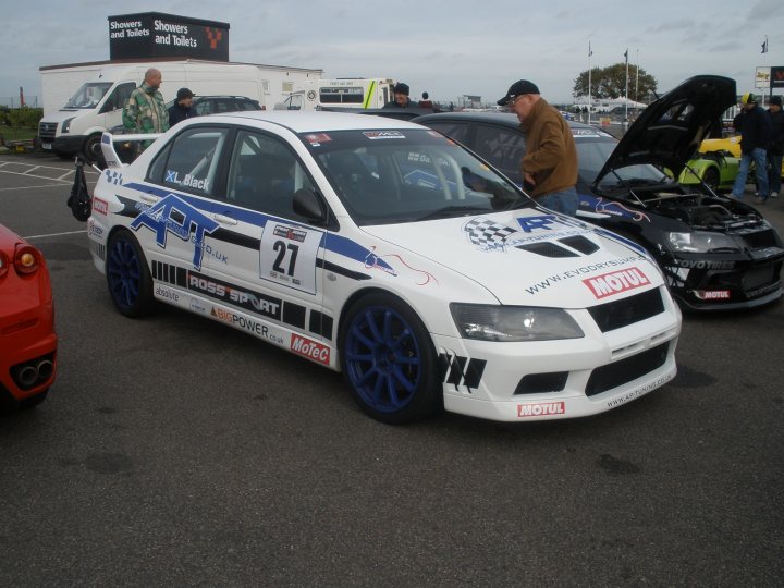 Thank you. - Page 1 - East Anglia - PistonHeads - The image displays a scene of car customization or racing. In the foreground, a white subcompact car with black accents is parked and features a number "21" on the side, along with sponsor logos. A few people are present, one close to the car potentially discussing or examining it, another slightly behind him, and three in the distance. The background includes more vehicles, indicating that this might be a parking lot or competition area for cars.