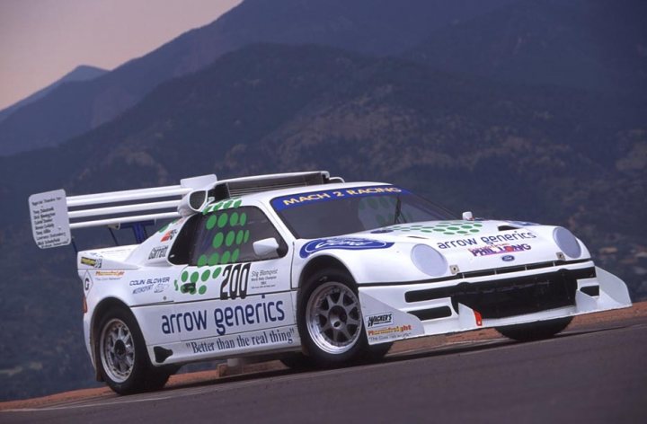 Businessman Rich Pistonheads Youre - The image features a Ford racing car that appears to be a prototype for a rally to Africa, as indicated by the "Arrows and Africa" name on the car. The car boasts sponsorship logos including those for "Panthers," "Fortaco," "Halfords," and "200," with the number "200" prominently displayed on the sidecar. The car has a distinctive dark color with a green and white camouflage pattern. It is seen racing on a road lined with gravel, set against a backdrop of rocky mountain terrain.