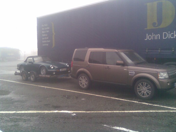 A truck that is sitting in the street - Pistonheads - The image depicts an outdoor scene where a flatbed truck is towing an open-top sports car. The truck has a flat-loading bed, and the sports car is positioned on top of its trailer, facing the same direction as the truck. The truck and car are situated on what appears to be a lot or road, with asphalt visible beneath them. The surrounding environment suggests cloudy weather conditions. There are texts and a logo visible with the words "John Dick" and an upper-case 'D' symbolizing a business association, but without additional context, their relevance to the scene is unclear.