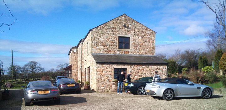 A Georgeous Run Out - Page 1 - Aston Martin - PistonHeads - The image showcases a man in a gray sweater and jeans standing between two parked cars, one black and one silver. He appears to be presenting or discussing the sports car, which is a white luxury vehicle. The setting is outdoors, in front of a farmhouse or barn-style building made of stone, with a clear blue sky overhead and well-maintained grass or lawn. The vehicles are neatly parked on what looks like a gravel or dirt road, suggesting a rural or semi-rural area.