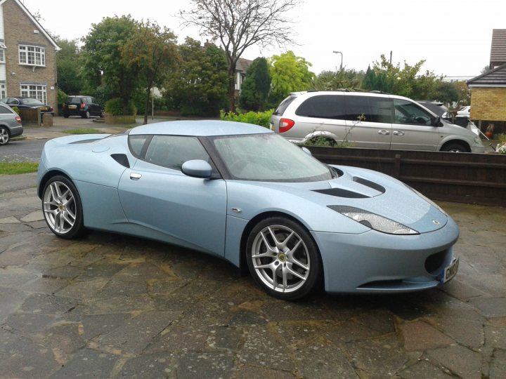 Picked up the Evora today  - Page 1 - Evora - PistonHeads - The image is a photograph showcasing a blue sports car parked on a partially visible driveway or road. The vehicle appears to be a convertible, characterized by its sleek design, a low and wide profile, and long hood. The car is equipped with alloy wheels and has visible emblems on the body, including one on the front and another on the side, indicating the model or brand. The car is parked facing away from the camera, with its rear end visible, and the driveway or road is lined with greenery and residential buildings. No people or distinctive landmarks are immediately noticeable, giving the photograph a calm and serene atmosphere.