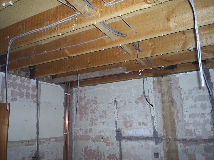 Our house renovation - lovely 1970's style decor - Page 1 - Homes, Gardens and DIY - PistonHeads - The image shows an unfinished interior space with exposed wooden studs forming the ceiling, visible insulation, and what appears to be electrical wiring hanging down. There is no drywall in place, revealing the structural framework of the room. The walls are being patched upwards from the ground, indicating renovation work. The ceiling structure crisscrosses with different length beams, and there is a visible hole in the upper right area. The space has a raw, factual look, absent of any decorative elements or finished surfaces. This is a photograph that captures the state of a room undergoing construction or renovation.