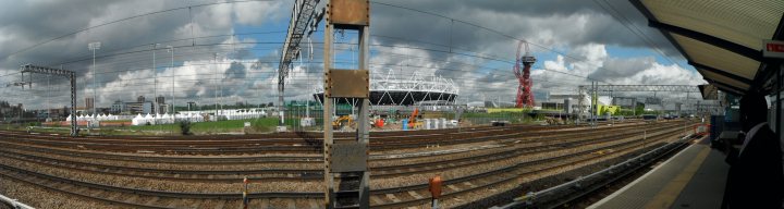 Visit to the Olympic Park - Page 1 - Sports - PistonHeads - The image captures a scene at train tracks on a cloudy day. Rows of train tracks extend towards the horizon, converging on a central location. Structures that could be trains or industrial equipment are present. Puffy gray clouds fill the sky, casting a muted light over the scene. The perspective creates a sense of depth and space, leading the viewer's eye towards the horizon.