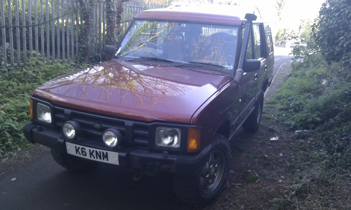 My new toy - Page 1 - Off Road - PistonHeads - The image features a maroon SUV parked on a dirt road. The vehicle is captured from the driver's side, with the headlights clearly visible and the side mirrors in the correct position. The registration plate is visible and reads "K6 KNM". The setting suggests a suburban or rural area, surrounded by lush greenery and partially obscured by large trees and bushes. The sky can be seen in the background, suggesting that the photo was taken during the day.