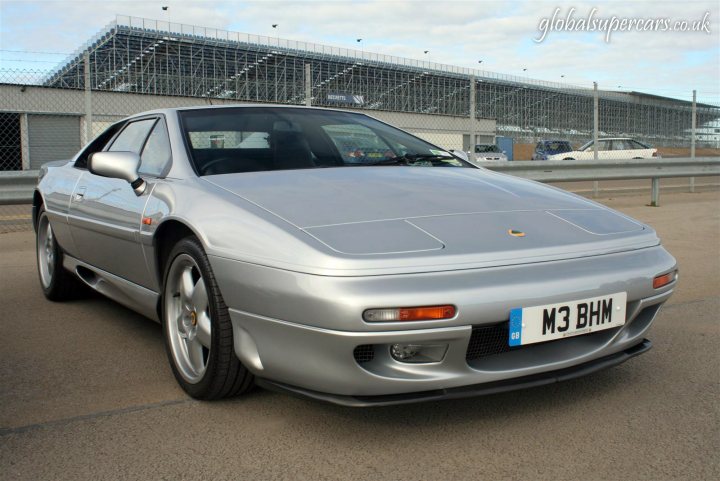 Snaps Pistonheads Silverstone Sunday Service Supply - The image showcases a silver sports car parked on a concrete surface, in front of a large metal structure suggesting a garage or workshop setting. The car features a distinctive design with round headlights and a bold front grille. It's equipped with alloy wheels, characteristic of sports cars, and the license plate reads "M3 BHM". The background is somewhat blurred, drawing attention to the car in the foreground.