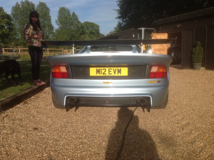 Another newbie - Page 1 - Noble - PistonHeads - In the image, a person stands on a stone surface behind a silver luxury car parked on gravel. The car, with its distinctive large rear spoiler, is parked in front of a wooden barn and a dog. The car's license plate shows the letters "MI2 EVM," and the person appears to be observing the vehicle. The overall setting suggests a rural or semi-rural environment.