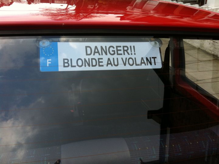 Peugeot 205 Blondemobile - Page 1 - Readers' Cars - PistonHeads - The image shows the back windscreen of a car with a sticker on it. The sticker is written in French and reads "DANGER! BLONDE AND DANGEROUS," translating to "Caution: Blonde and Dangerous" in English. The car is parked, and the background is blurred, suggesting the focus is on the sticker. The sticker appears to be handmade, with a playful and humorous intent rather than a serious warning. This type of sticker can be often seen as a form of humor in some countries.