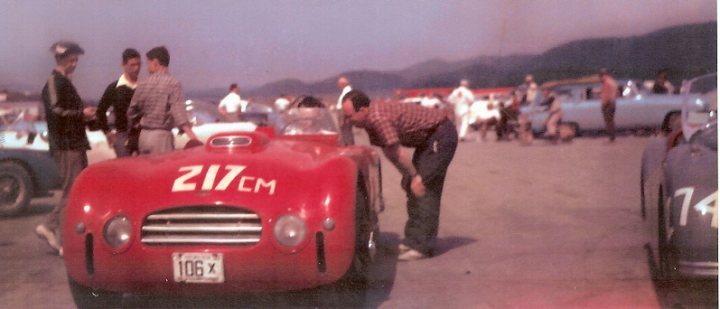 Early TVR Pictures - Page 33 - Classics - PistonHeads - This is a color photograph that captures a moment of interaction between various groups of people and vintage cars. In the foreground, there is an old-fashioned red car labeled with numbers and letters on its front and side. A man is shown examining the vehicle from a kneeling position, while other individuals stand nearby, engaged in conversation. The cars in the background suggest a gathering or event, possibly featuring antique automobiles. The environment is outdoors, with the vehicles parked on a flat surface, and there appears to be a mountainous landscape in the distance.