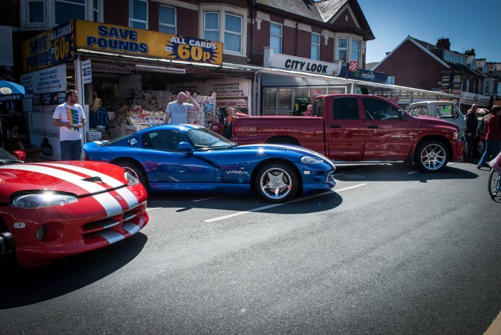 A car that is sitting in the street - Pistonheads - The image depicts a city street scene featuring a few vintage cars parked along the curb. The street is lined with storefronts, and it appears to be a populated shopping district. There's a variety of vehicles, including a pair of red cars, one of which is a vintage Dodge Viper parked in front of a blue car, and another red car parked behind it. A group of people, possibly interested in the cars or passing pedestrians, are scattered throughout the scene, adding to the liveliness of the urban setting.