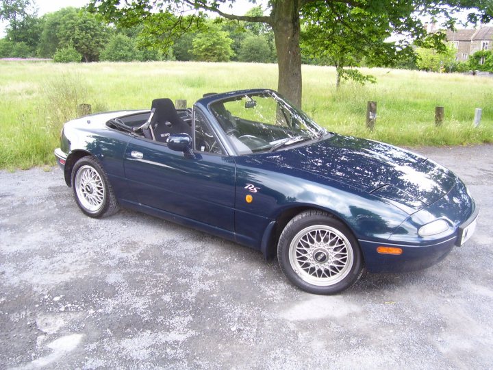 Pistonheads - The image depicts a blue Mazda Miata sports car parked on a concrete surface, likely in a driveway or parking lot. The car is a two-door convertible, featuring a silver top that is currently down. It has a textured silver hood and consistent blue bodywork. There are trees in the background, suggesting this is a suburban or rural setting. The tires have a white wall design, adding to the vintage aesthetic of the car.