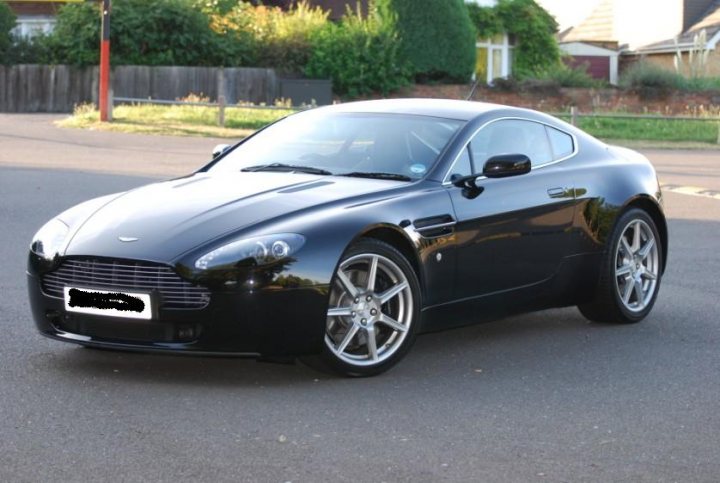 Pistonheads Promised - The image displays a sleek black Aston Martin sports car viewed from a rear side angle. It is parked on an asphalt surface under bright light. The car features a prominent front grille and sculpted lines typical of luxury sports cars. The construction indicates it is parked in a suburban area with houses and shrubbery in the background, though the focus is on the vehicle. There is no visible text in the image to provide context or additional information.