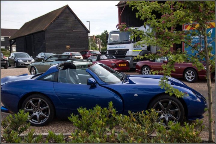 TVRCC + Guests Berkshire June Meet - Page 1 - Thames Valley & Surrey - PistonHeads - The image showcases a vibrant setting, featuring a striking blue convertible sports car as the focal point. The sports car is parked on a gravel area, surrounded by several other cars, trucks, and a trailor, indicating a well-used parking lot. A small white building with a wooden roof forms the background, adding to the rustic charm of the scene. A few trees and bushes can be seen, suggesting the presence of natural elements. A blue sky, visible in the upper left corner of the image, completes the picturesque setting.