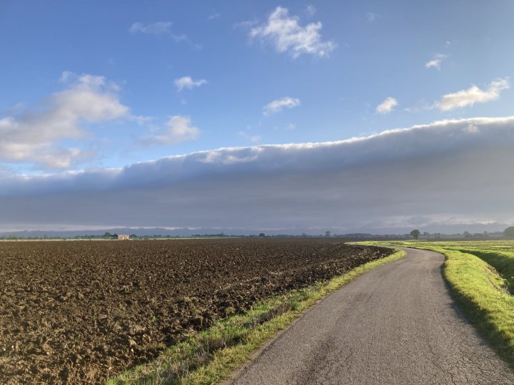 The "Photos From Today's Ride" thread... - Page 471 - Pedal Powered - PistonHeads - The image portrays a serene rural scene. A paved road, possibly a farm track, cuts through the landscape, leading towards a vast field that stretches out into the horizon. The field is currently plowed, with the soil appearing in neat rows. On either side of the road, there are areas of green vegetation and open land. Above, a sky filled with clouds spans across the scene, suggesting an overcast day. The overall mood of the image is peaceful and untouched by urban development.