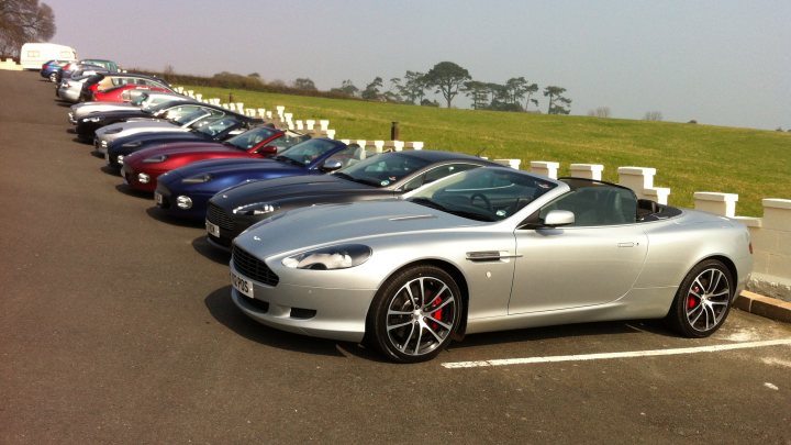 South west breakfast club - Page 1 - Aston Martin - PistonHeads - The image depicts a neat and orderly car show on a grassy hill, with a long row of luxury cars, all painted in a variety of colors, displayed next to each other. The cars range from different makes and models, visible in the array of front ends, adding to the diversity and appeal of the show. The cars are parked diagonally, both across and along the row, creating a semi-circle around the viewing area. The backdrop features a beautiful open sky and lyrical trees, enhancing the aesthetics of the scene.