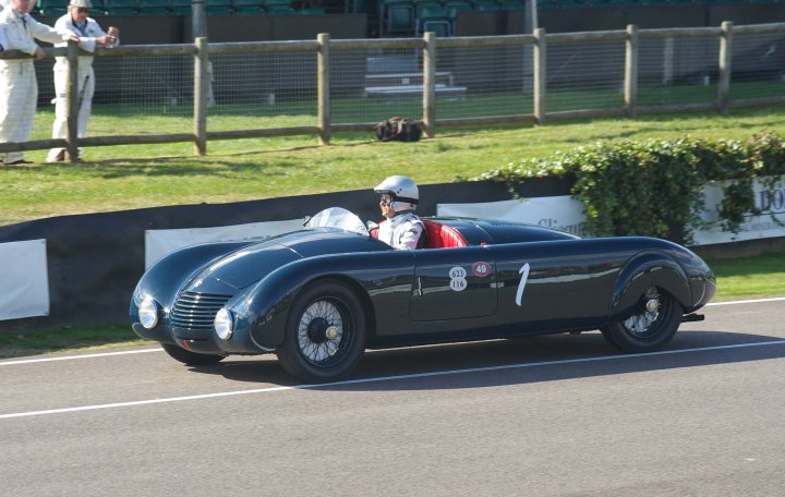 2012 Goodwood Revival Meeting Photo Gallery - Page 1 - Goodwood Events - PistonHeads - The image captures an intense moment of a vintage racing car zooming around a track. The car, painted in a sleek black color, is captured mid-track with its driver, donned in racing gear, holding the steering wheel with both hands. The track, made of asphalt, stretches out in the background with a grassy shoulder, adding a touch of nature to the otherwise adrenaline-fueled scene. A wooden fence and chairs can also be seen, perhaps indicating a viewing area for spectators. The overall scene is charged with the excitement and adrenaline associated with motor racing.