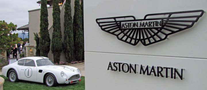 Pics from Aston event, Monterey - Page 1 - Aston Martin - PistonHeads - The image captures an outdoor scene showcasing a classic white sports car on a grassy area, placed in front of a wall with a well-lit logo that reads "ASTON MARTIN". The logo is prominent and features the classic Aston Martin wings design. The sports car, despite appearing somewhat aged, retains an air of elegance and sophistication. The setting suggests a possible dealership or event related to the automotive brand.