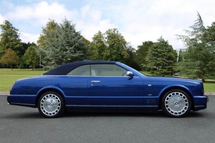 A car with a surfboard strapped to the back - Pistonheads - The image shows a sleek blue convertible parked on a driveway. The car, appearing to be in excellent condition, is positioned at an angle towards the camera. The vehicle has distinctive wheels, and the window is slightly open. In the background, there are tree-filled greenery and a glimpse of an open field. The sky is partly cloudy, suggesting a pleasant day. The overall scene exudes a sense of tranquility and luxury.