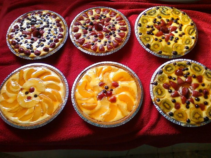 The image showcases six individual pies arranged on a red cloth. Each pie is encased in a tin foil wrapper, presenting a sesame seed coated top crust. The colorful filling reveals a mix of yellow, orange, and red, likely from strawberries and cherries, suggesting a fruit-based tart filling. From left to right, the pies exhibit slightly varied toppings of the fruits, adding to their visual appeal.