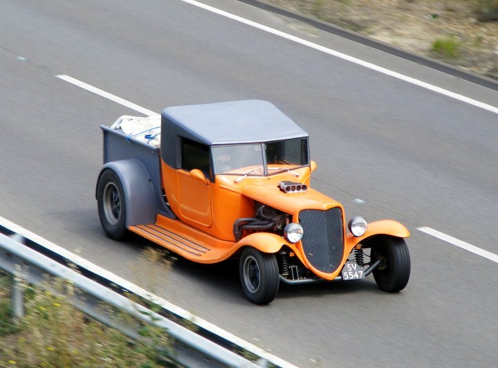 A red truck parked on the side of a road - Pistonheads - The image showcases a small orange car-like vehicle driving on a two-lane road. It is a picturesque moment, likely on a sunny day, with the vibrant color of the vehicle standing out against the neutral tones of the road. The road itself appears to be in good condition, with white lines indicating divide markers for separate lanes of traffic. There are high grass and shrubs alongside the road, suggesting a rural or suburban setting. There is no visible traffic, giving the scene a peaceful and calm atmosphere.