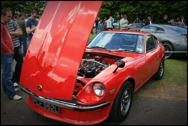 Pistonheads Tatton Park Classic - The image features a vibrant red sports car with its hood propped open, revealing the engine beneath. The car is parked on a grassy field, with the hood gently supported by a rod. In the background, there's a crowd of people gathering, suggesting that this might be a car show or an informal gathering of car enthusiasts. The car is compact yet seems sturdy, with its sleek design and powerful engine. The scene conveys a sense of anticipation and admiration for the vehicle.