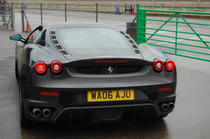 Combe Castle Supercar Pistonheads Stroke Association Day - In the image, there is a black Ferrari sports car parked on the side of a street. The car is positioned at a slight angle to the camera, showcasing the front and side of the vehicle. The license plate of the car is clearly visible and reads "W06 AJU". The car is stationed behind a green fence, providing a vibrant contrast to the sleek black color of the Ferrari. In the background, there are stadium seats visible, indicating that this scene might be taking place near a sports arena or similar venue.