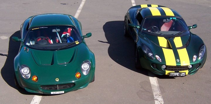 Show us your hard top - Page 1 - Elise/Exige/Europa/340R - PistonHeads - The image features two sports cars parked side by side on a tarmac. The cars are green with yellow stripes on the roof and hood, and they both display the brand Lotus on the side. The car on the left angles its view across the lot when viewed from the driver's seat, while the car on the right is aligned with the camera, appearing to be parked perpendicular to the line. Both cars have black roofs and their passenger headrests are visible. The setting suggests a busy parking lot with clear skies overhead.